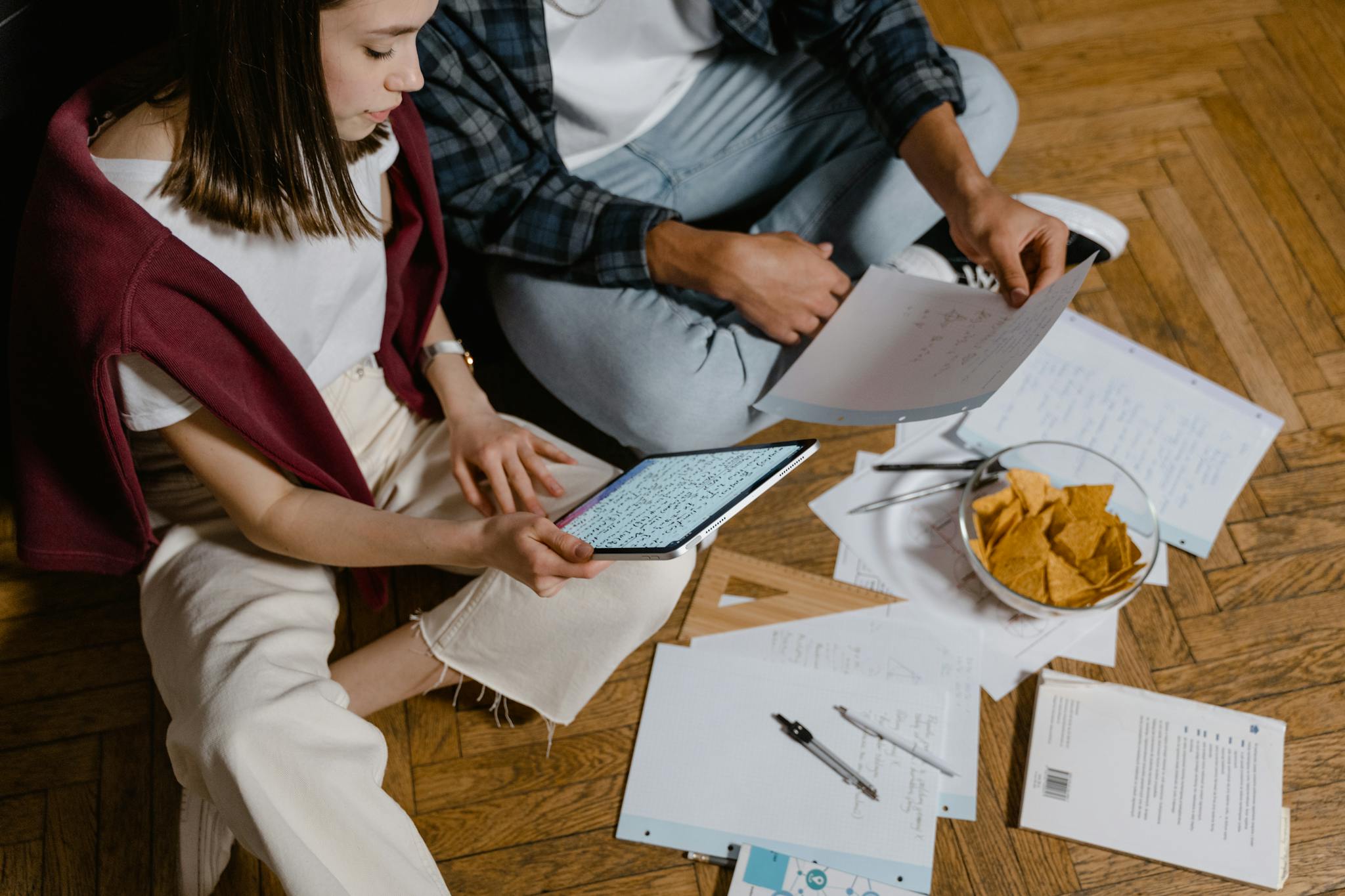 A man and woman study together on a wooden floor using technology and papers.